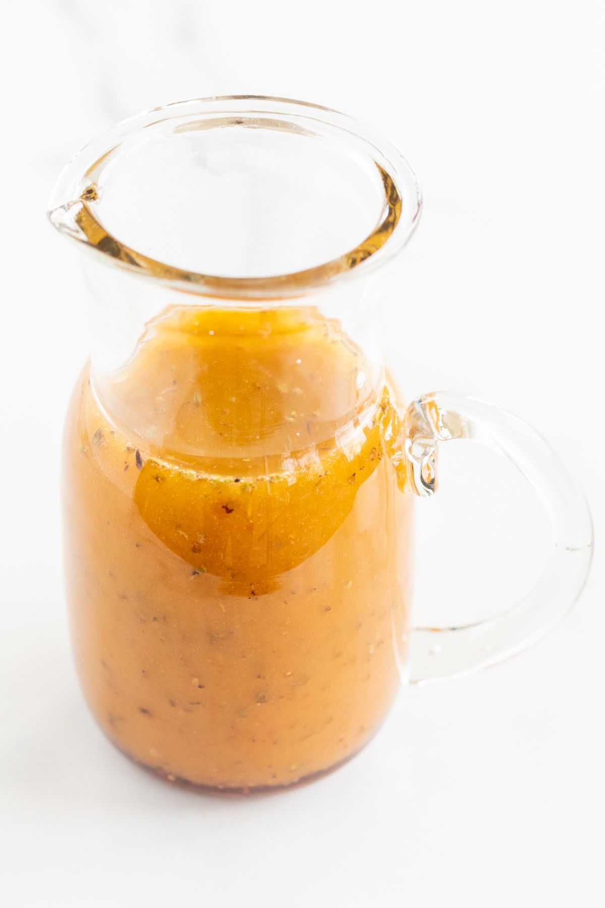 A small glass pitcher full of homemade Greek Salad dressing, on a white countertop.