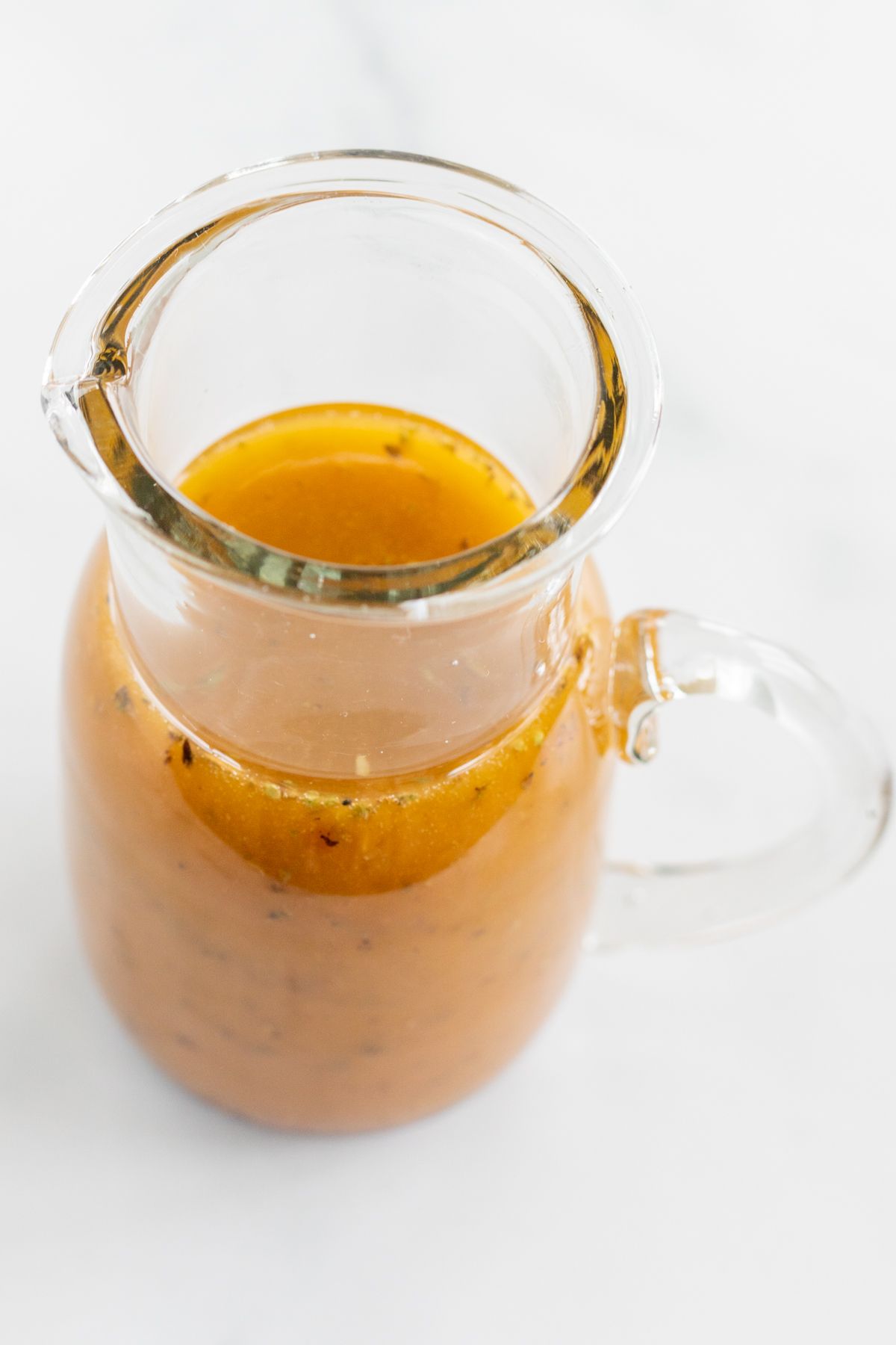 A small glass pitcher full of homemade Greek Salad dressing, on a white countertop.