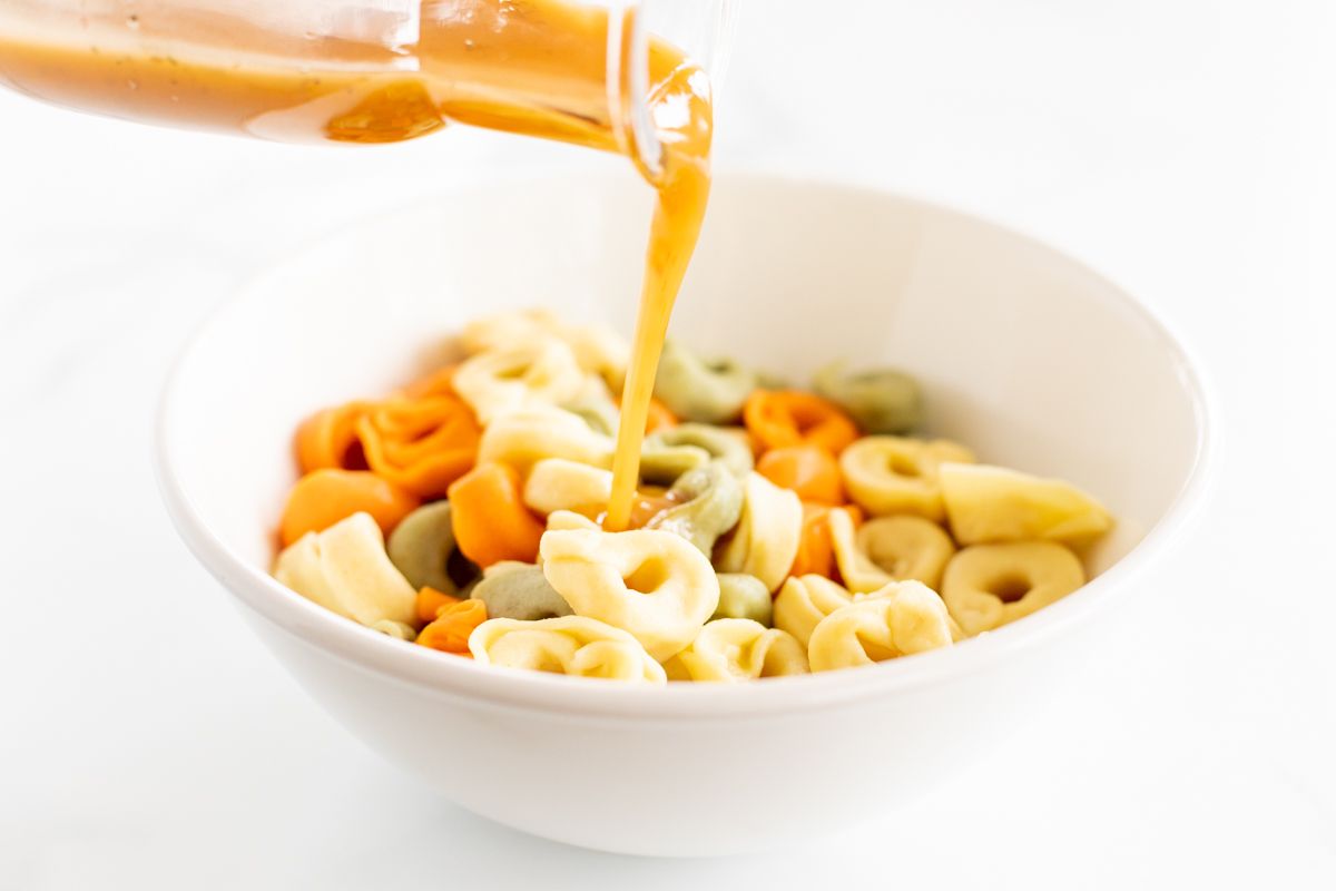 Greek salad dressing being poured over a white bowl full of stuffed pasta for a pasta salad recipe.