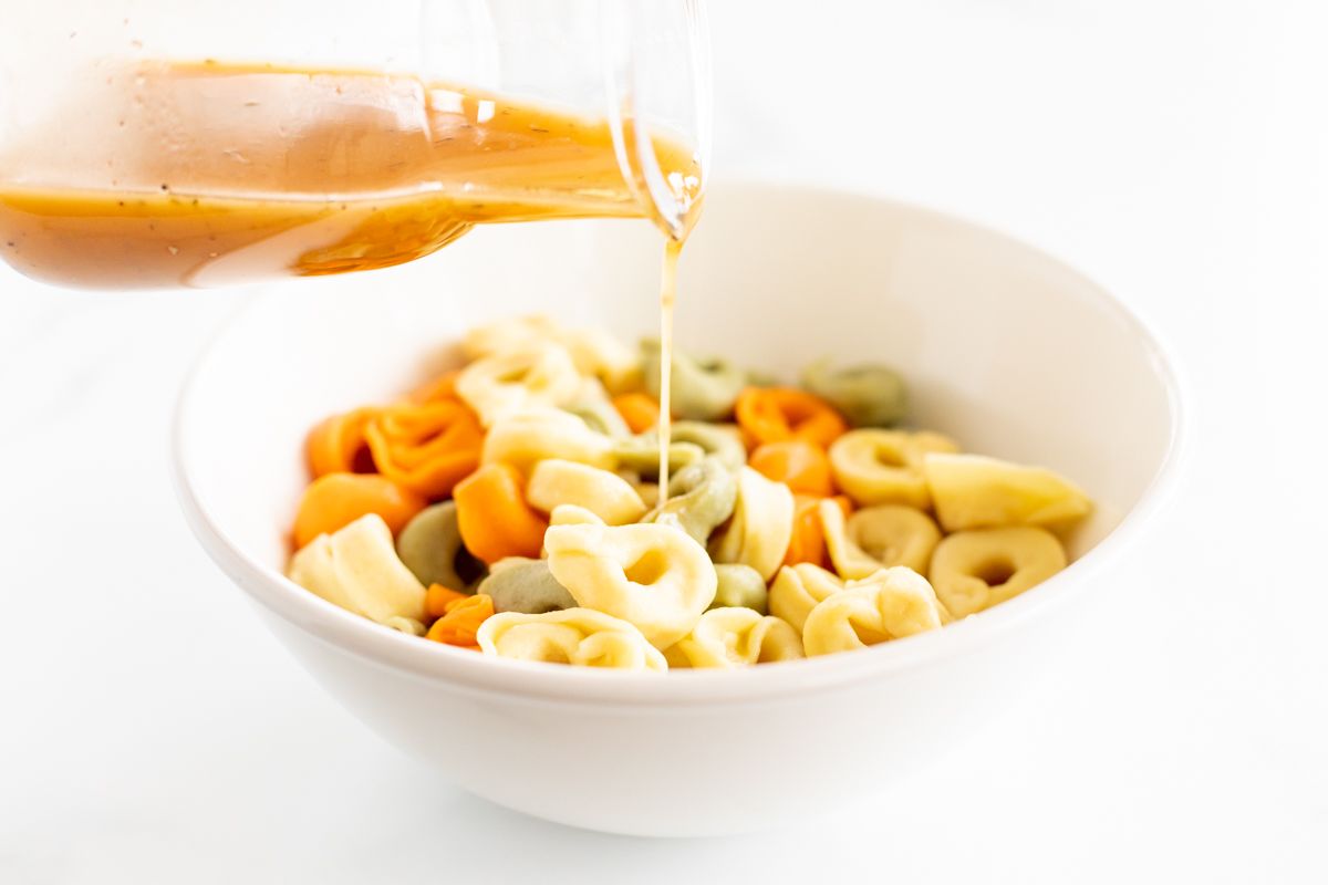 Greek salad dressing being poured over a white bowl full of stuffed pasta for a pasta salad recipe.