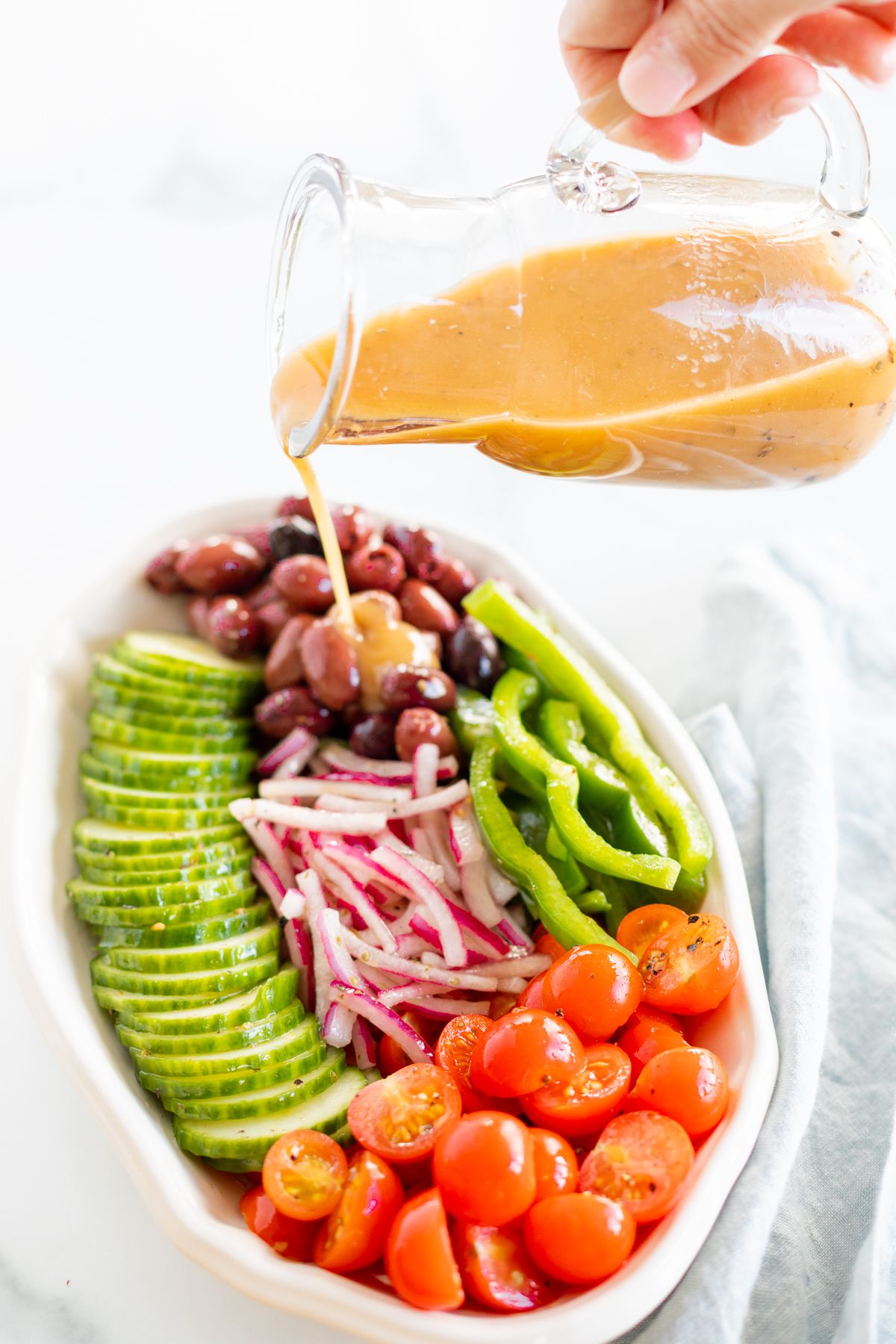 A small glass jar of Greek salad dressing pouring over a colorful Greek Salad on a white platter. 