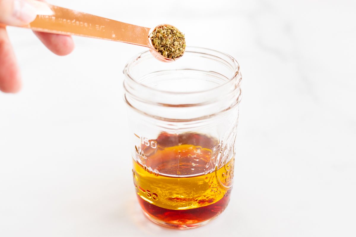 Ingredients for a Greek salad dressing recipe being added to a glass jar.