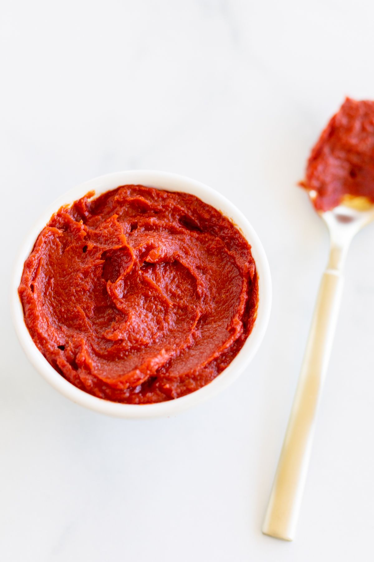 A small bowl of tomato paste on a marble countertop, gold spoon full of tomato paste substitute to the side.