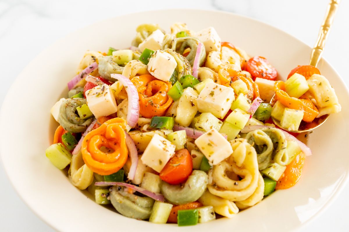 A white serving bowl full of tortellini pasta salad, gold serving spoon on the side.