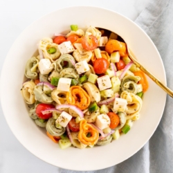 A white serving bowl full of tortellini pasta salad, gold serving spoon on the side.