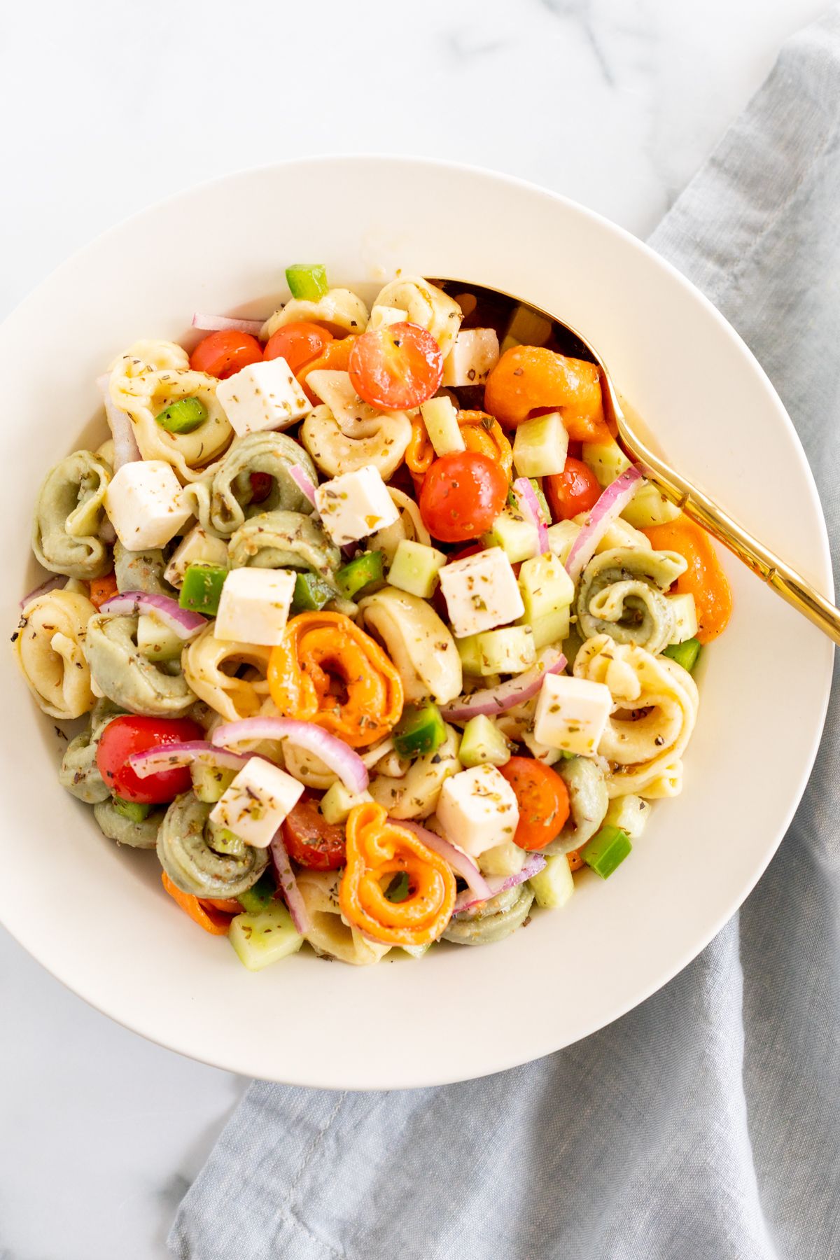 A white serving bowl full of tortellini pasta salad, gold serving spoon on the side.