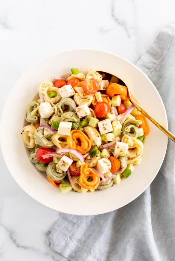 A white serving bowl full of tortellini pasta salad, gold serving spoon on the side.