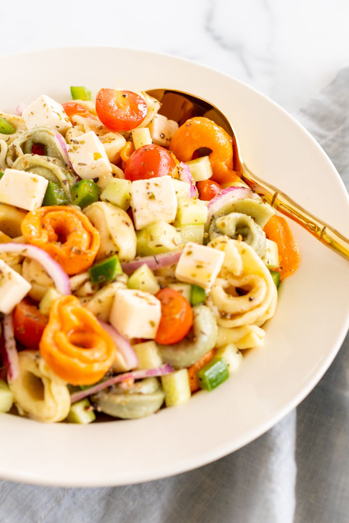 A white serving bowl full of tortellini pasta salad, gold serving spoon on the side.