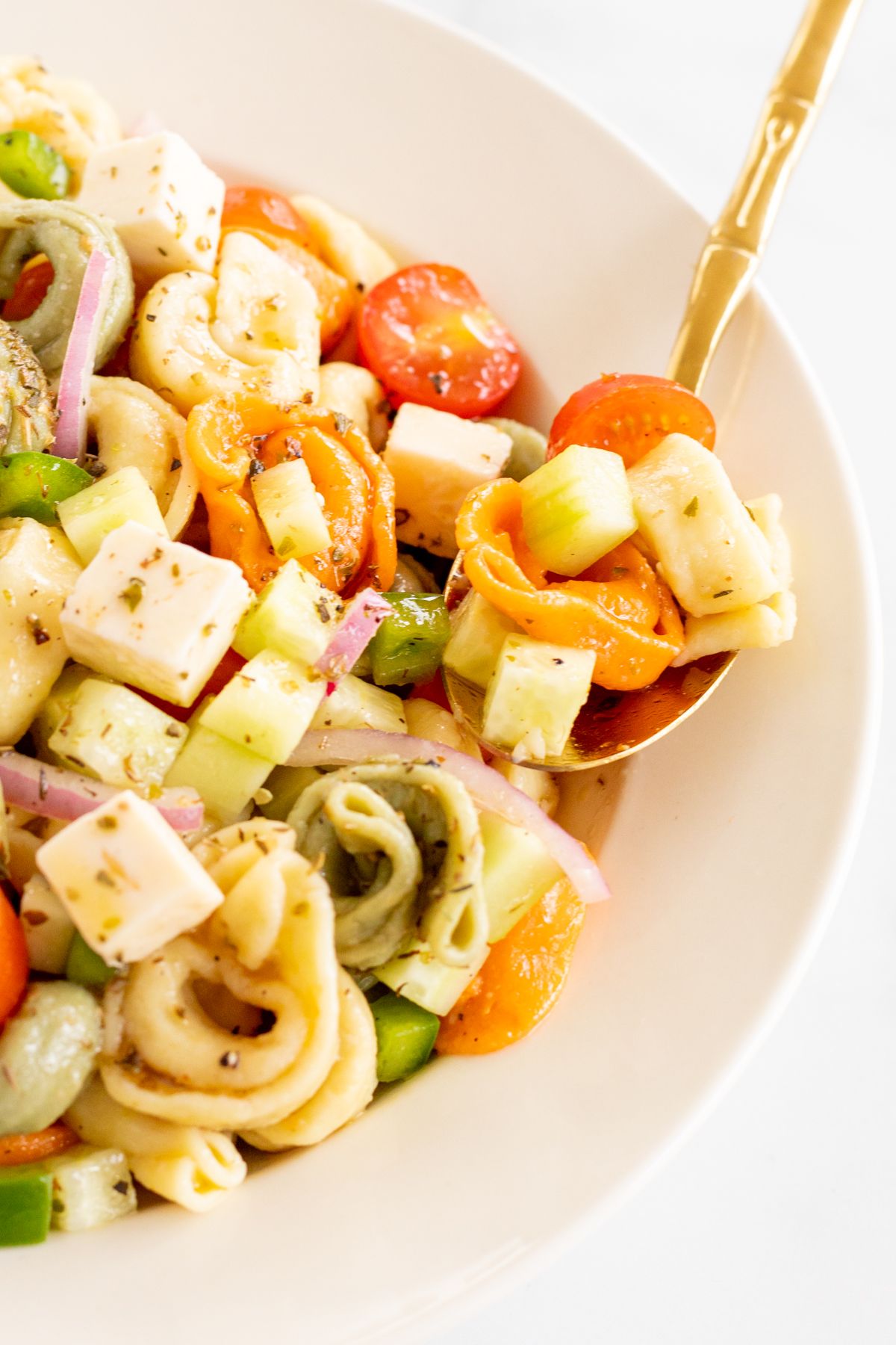 A white serving bowl full of tortellini pasta salad, gold serving spoon on the side.