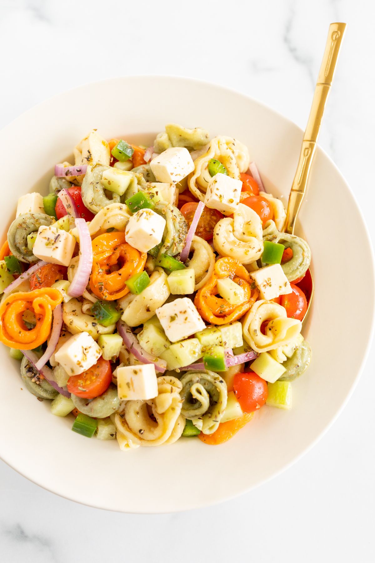 A white serving bowl full of tortellini pasta salad, gold serving spoon on the side.