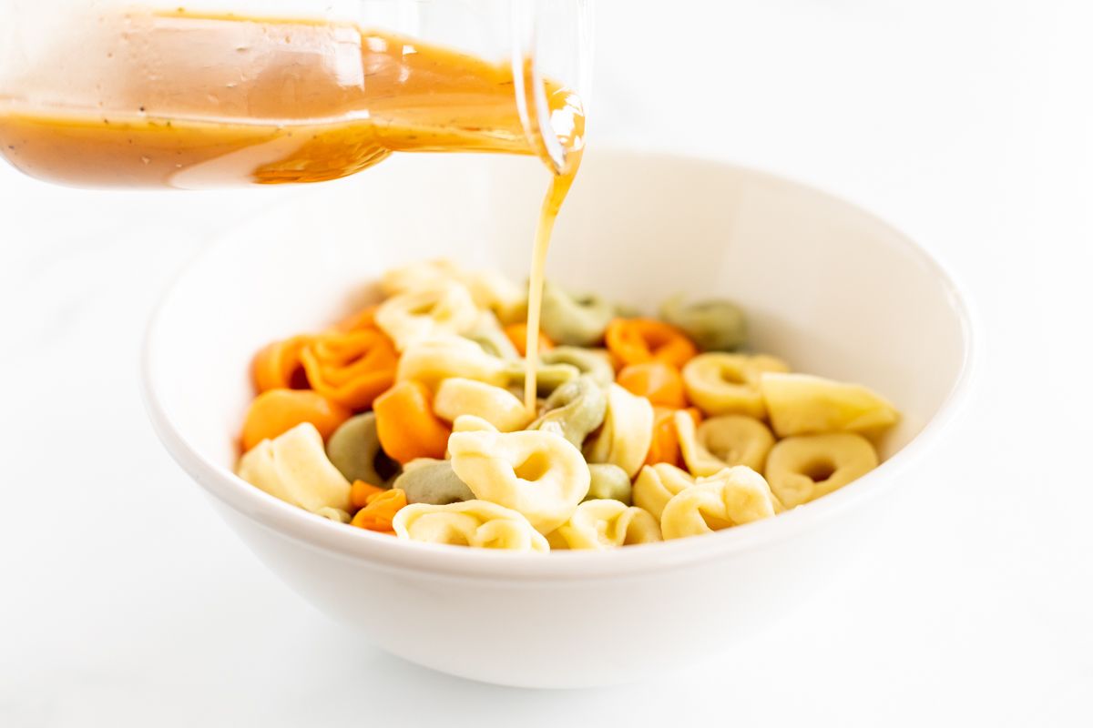 A bottle of homemade dressing pouring over a white bowl full of tortellini.