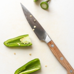 Overhead view of jalapeno peppers on cutting board with paring knife; one pepper has top cut off, the other is cut in half lengthwise