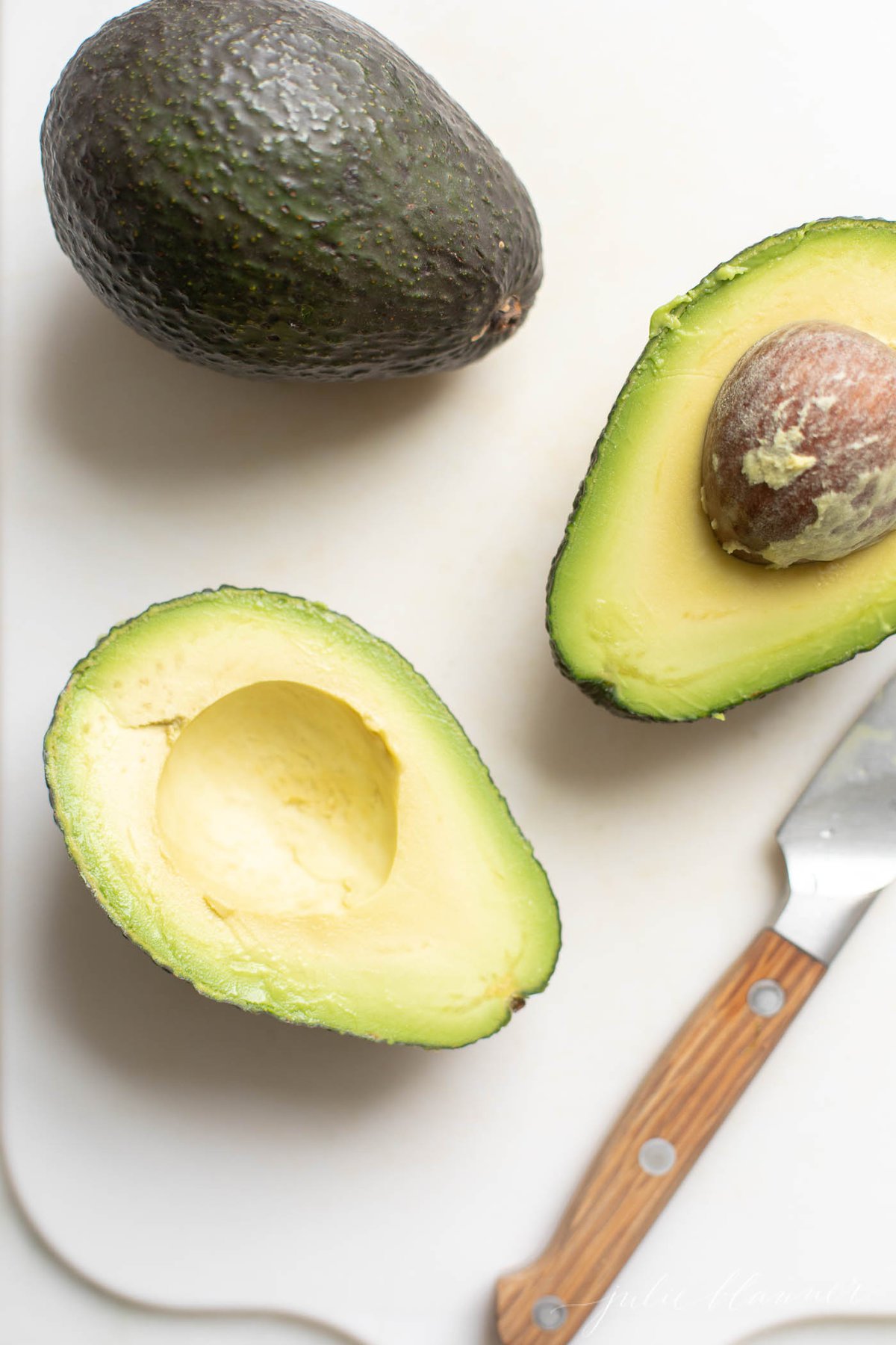 Overhead view of one whole avocado and two cut avocado halves with knife on white cutting board
