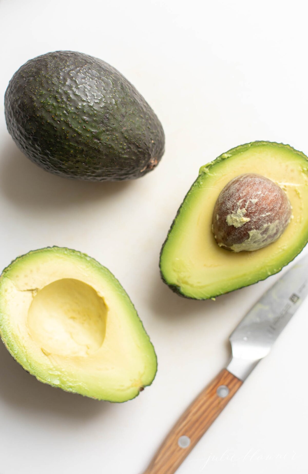 Overhead view of one whole avocado and two cut avocado halves with knife