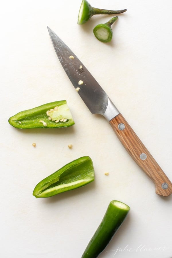 Overhead view of jalapeno peppers on cutting board with paring knife; one pepper has top cut off, the other is cut in half lengthwise