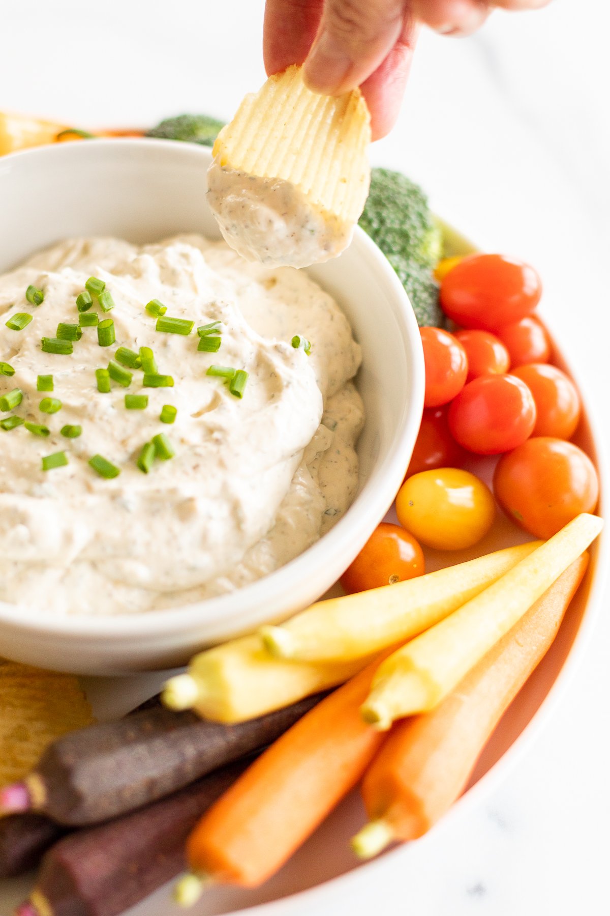 Lifting ridged potato chip from ranch dip in bowl set on serving platter with veggies