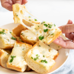 Bowl of garlic cheese bread set on pale blue linen cloth, hands pulling apart a cheesy slice.