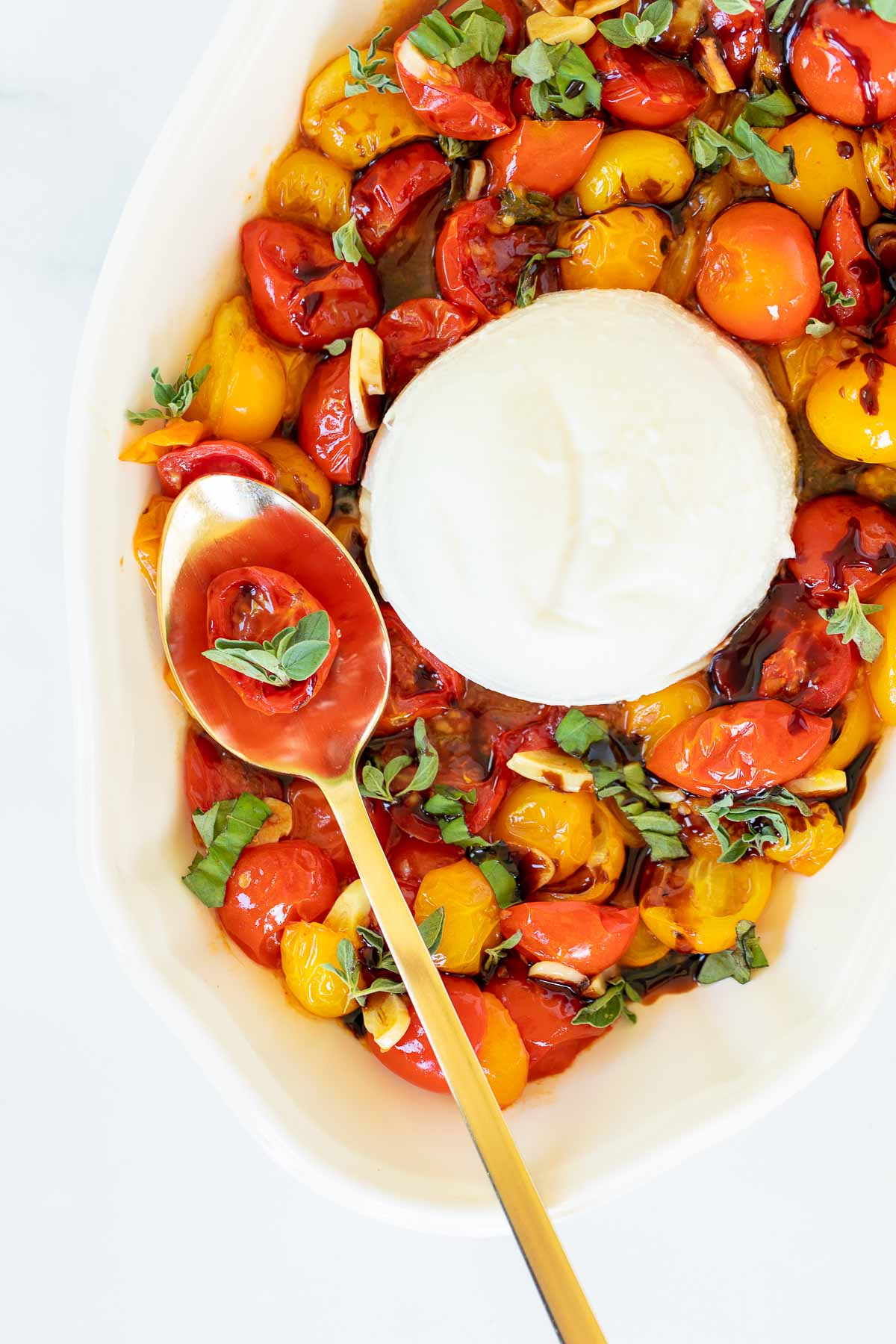 Overhead view of blistered tomatoes on serving platter with burrata, with single tomato and basil leaves on spoon