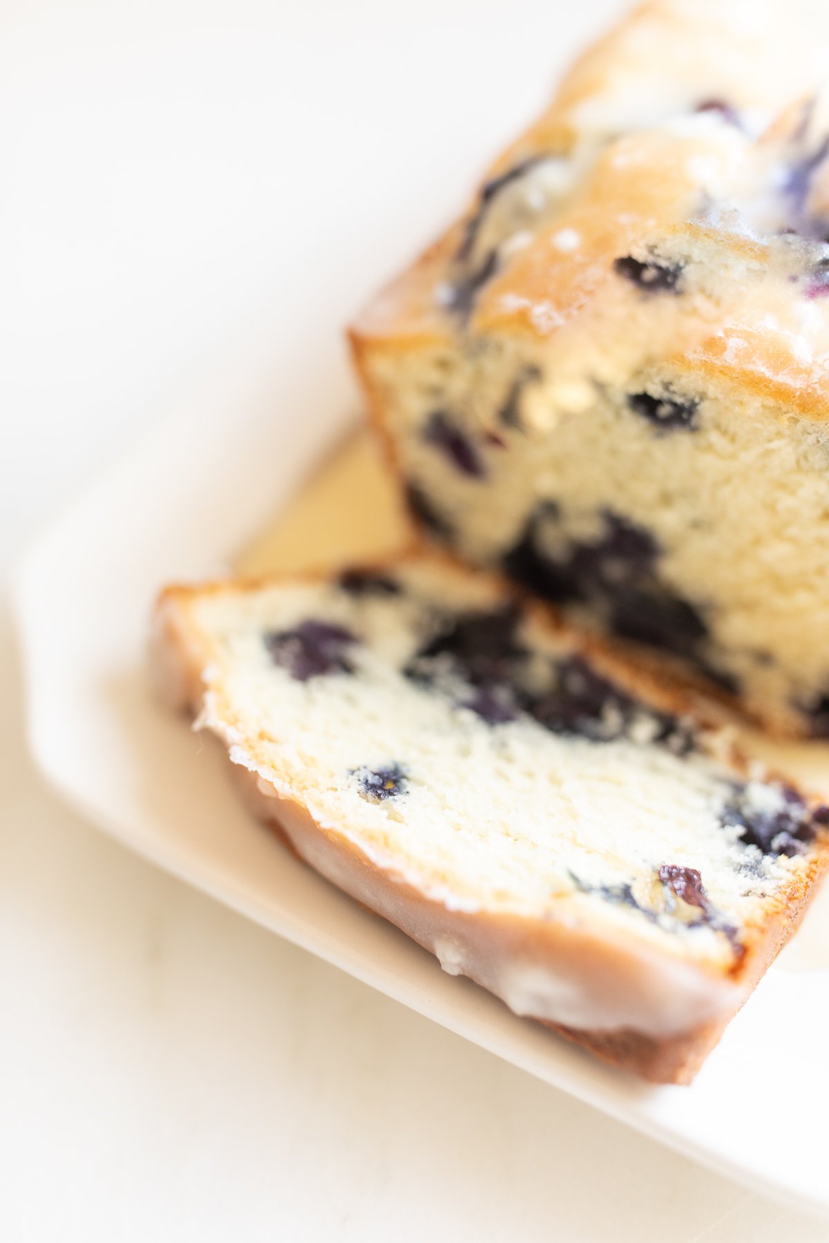 A loaf of blueberry bread, topped with a glaze. One slice is cut at the end, and it's on a white platter.