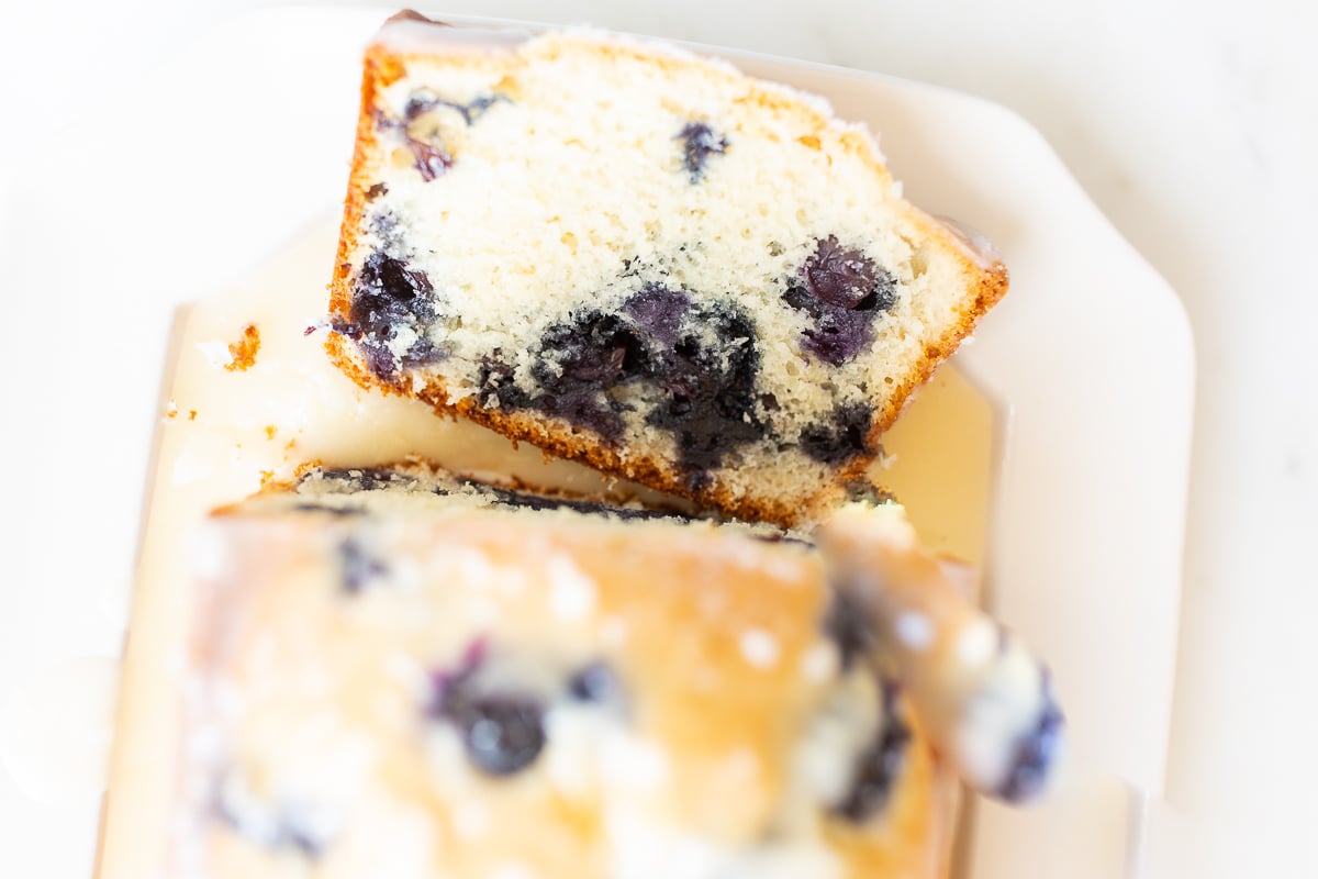 A loaf of blueberry bread, topped with a glaze. One slice is cut at the end, and it's on a white platter.