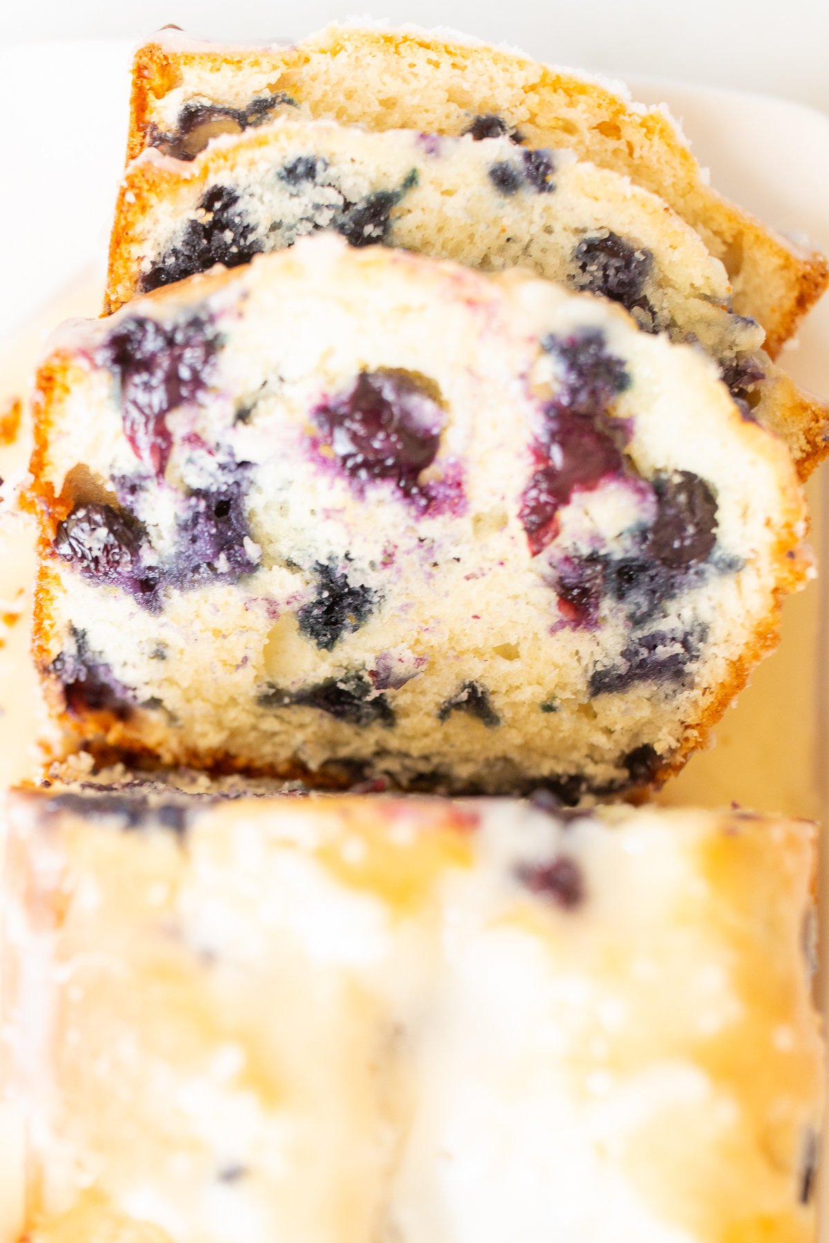 A loaf of blueberry bread, topped with a glaze. Two slices are cut at the end, and it's on a white platter.