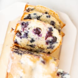 A loaf of blueberry bread, topped with a glaze. Two slices are cut at the end, and it's on a white platter.