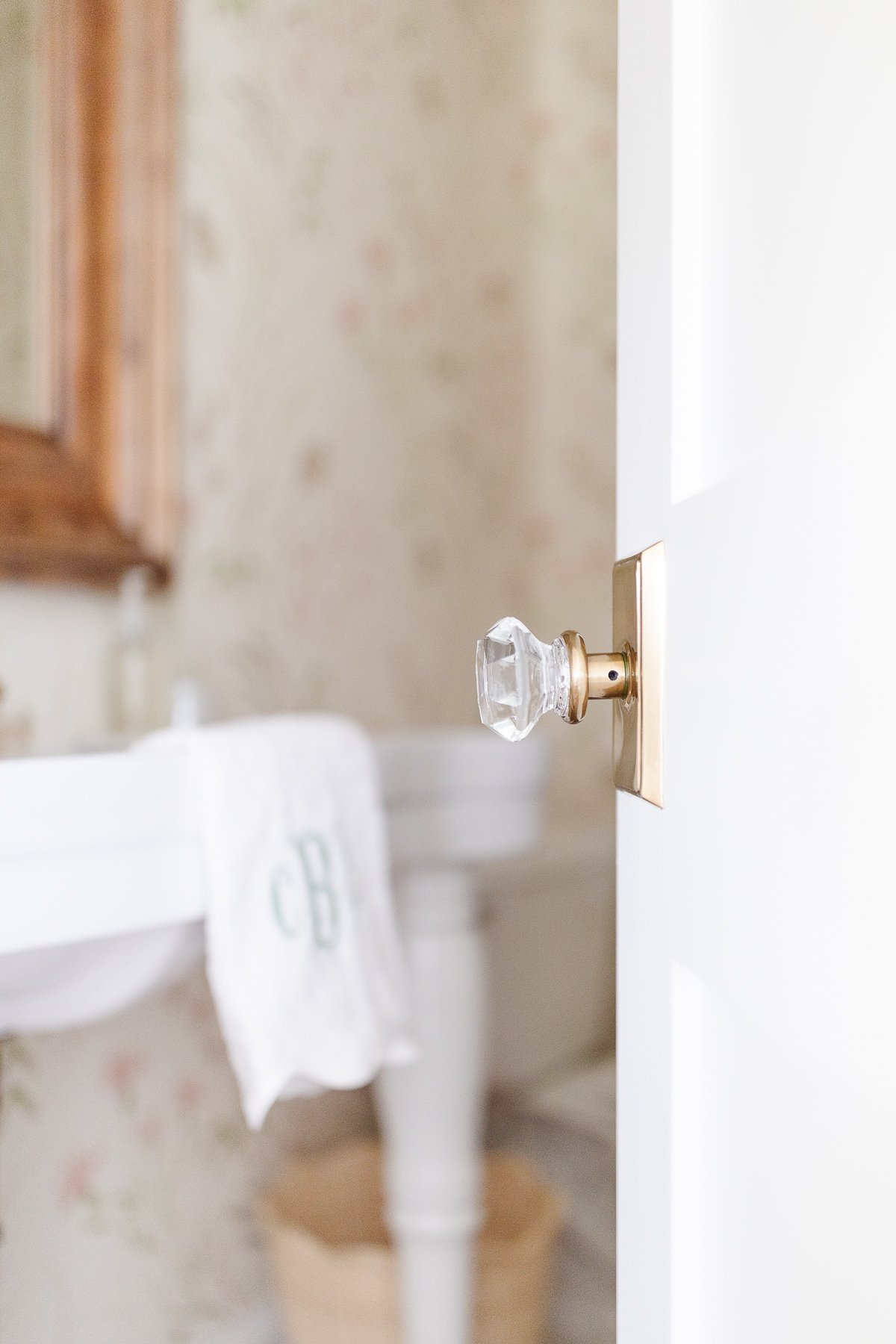 A brass door knob on a white door, partially open to reveal a bathroom with floral wallpaper and a hanging towel embroidered with initials.