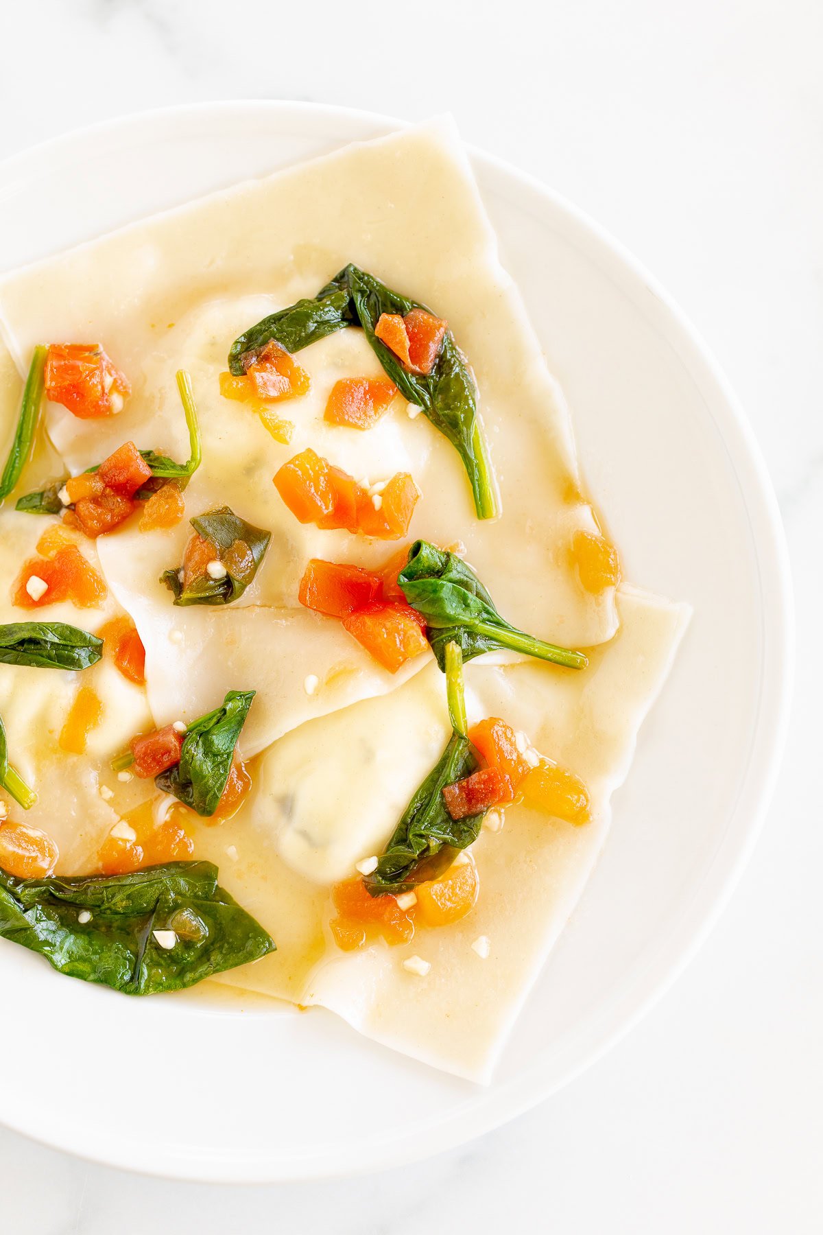 A plate of goat cheese ravioli topped with spinach and chopped tomatoes, served on a white marble surface.