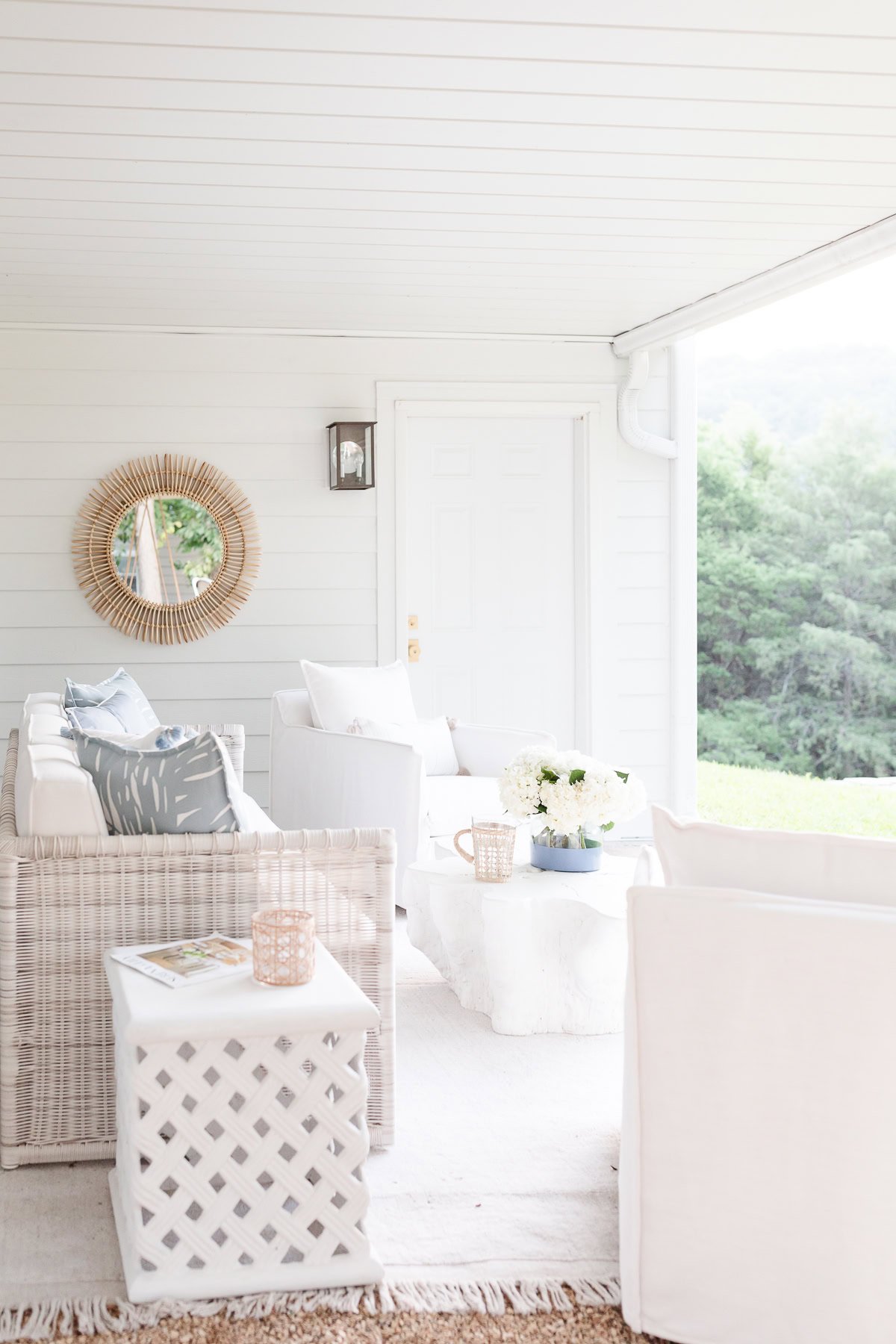 A sunlit porch with white furniture and pillow inserts, a lattice side table, and a round mirror on a light wooden wall. A bouquet of white flowers and decorative items are on the coffee table.