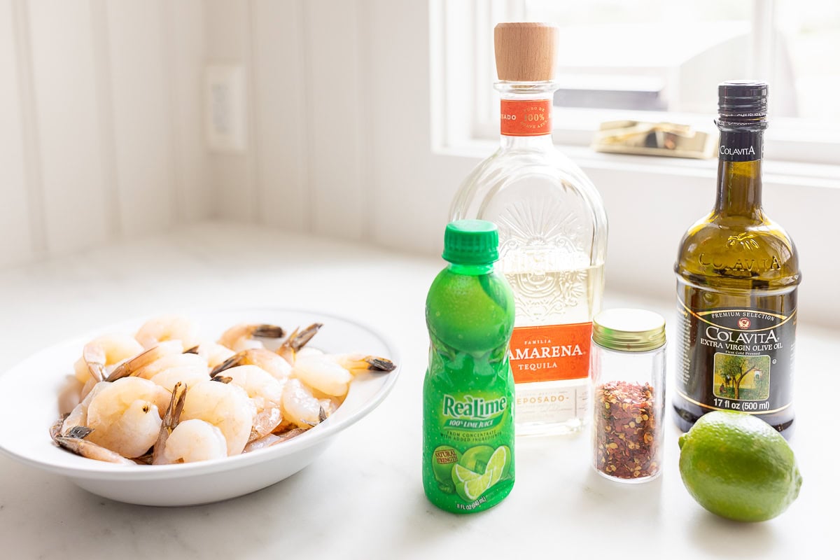 A kitchen countertop holds a bowl of tequila lime shrimp, a bottle of olive oil, lime juice, a fresh lime, and red pepper flakes.