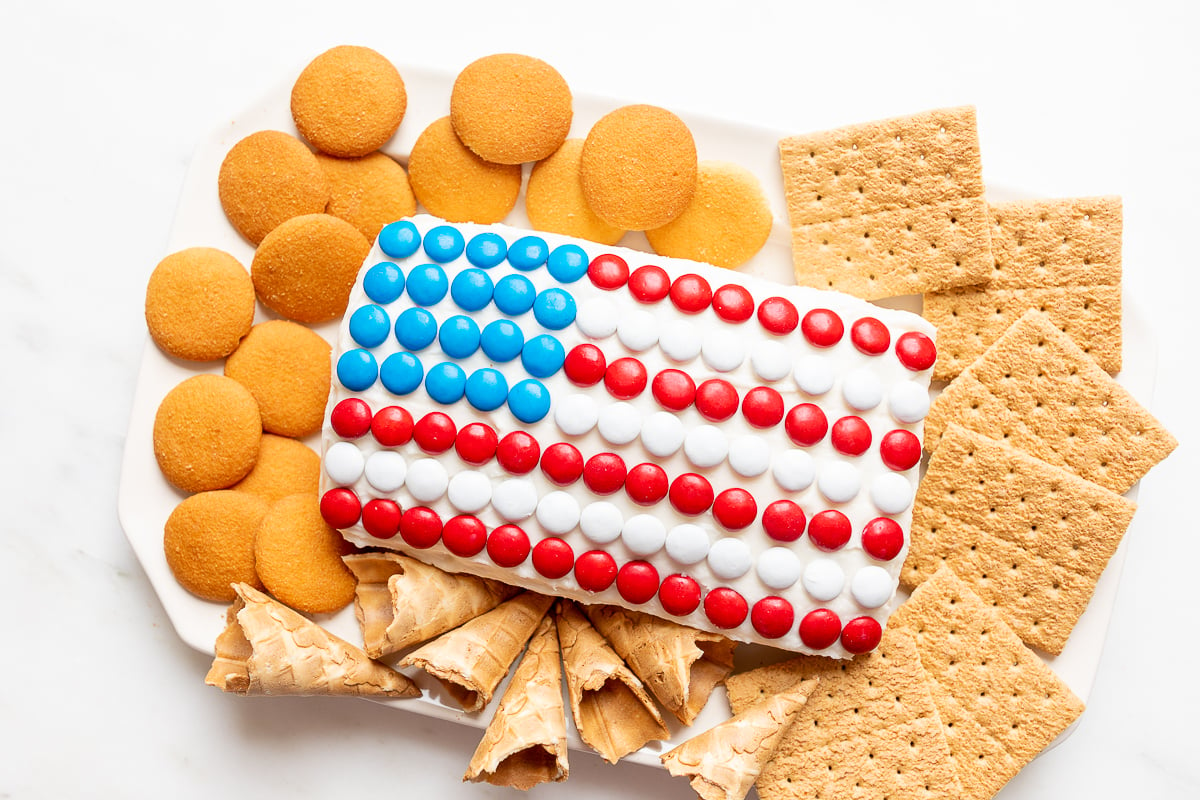 A red white and blue snack made of Funfetti dip in a flag shape, on a plate of cookies and crackers.