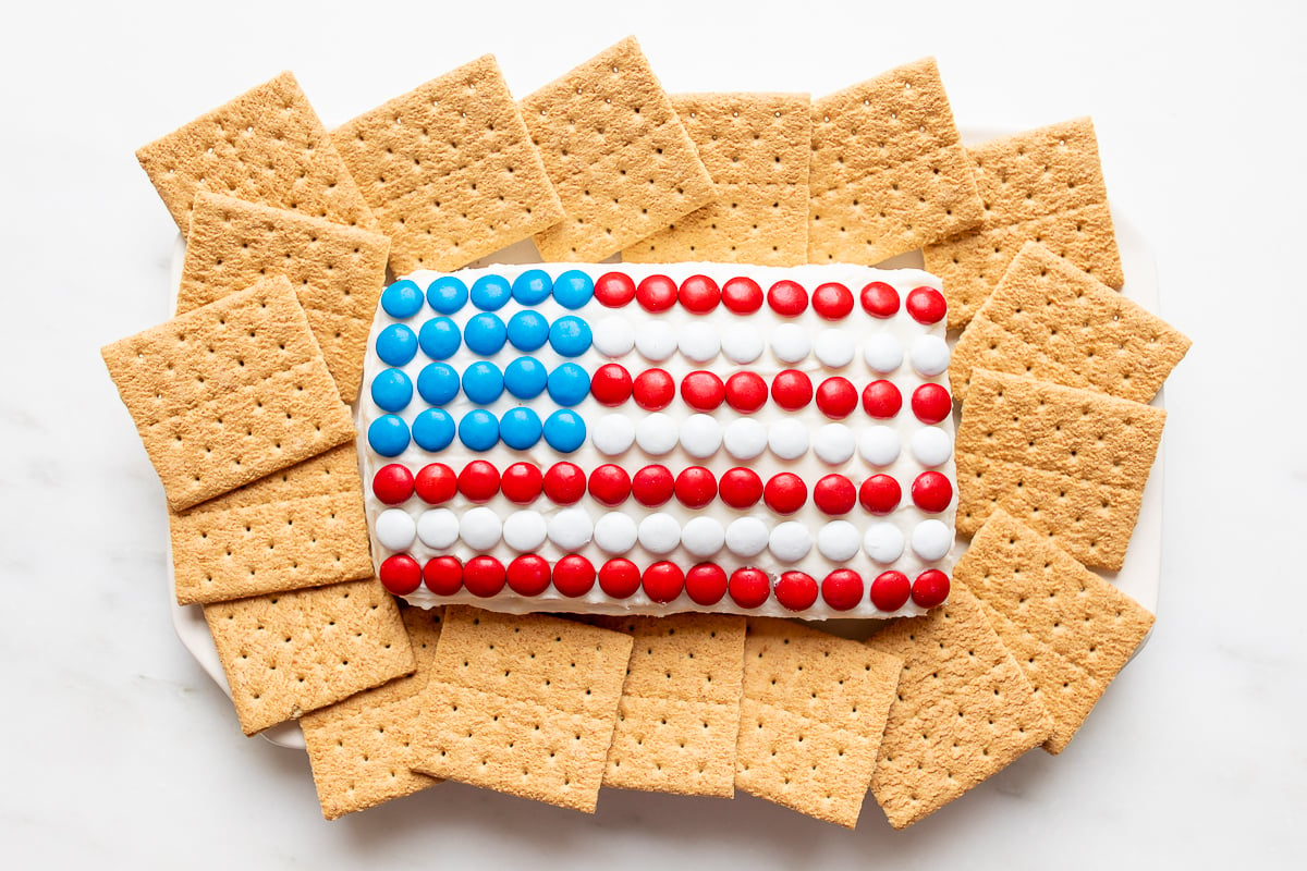 A red white and blue snack made of Funfetti dip in a flag shape, on a plate of cookies and crackers.