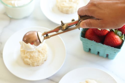 A biscuit topped with whipped cream and a box of strawberries in the background for a strawberry shortcake recipe.