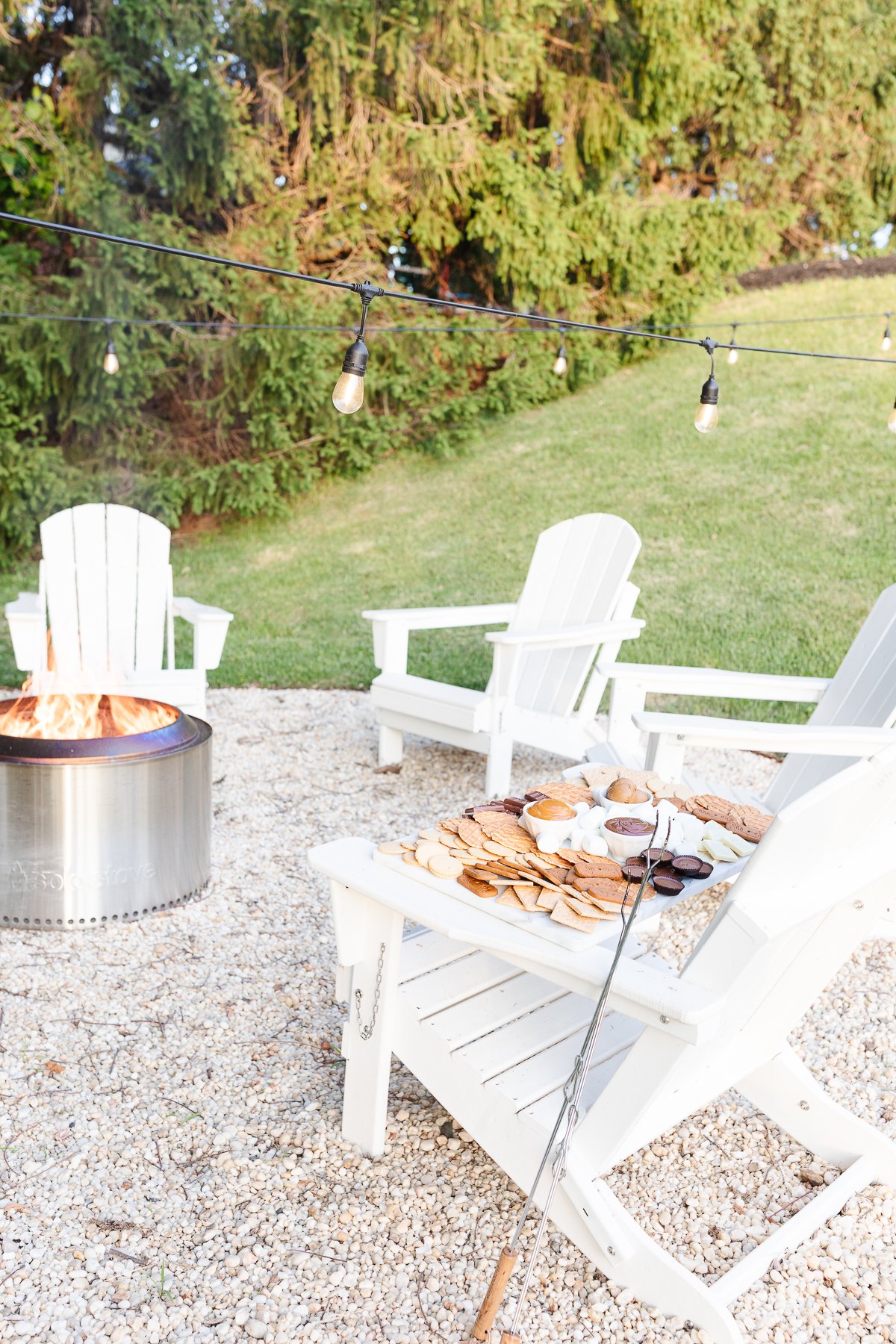 A Solo stove campfire on a gravel fire pit, surrounded by white chairs. A smores charcuterie board rests on one chair.