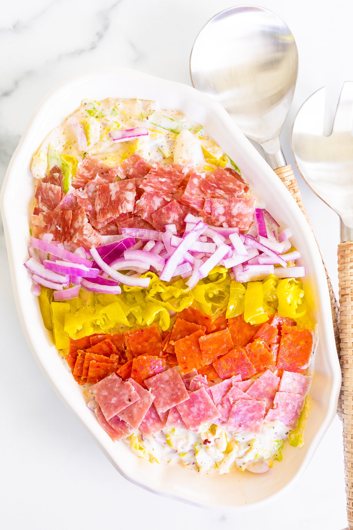 A grinder salad recipe in a white oval bowl, with rattan serving utensils to the side.