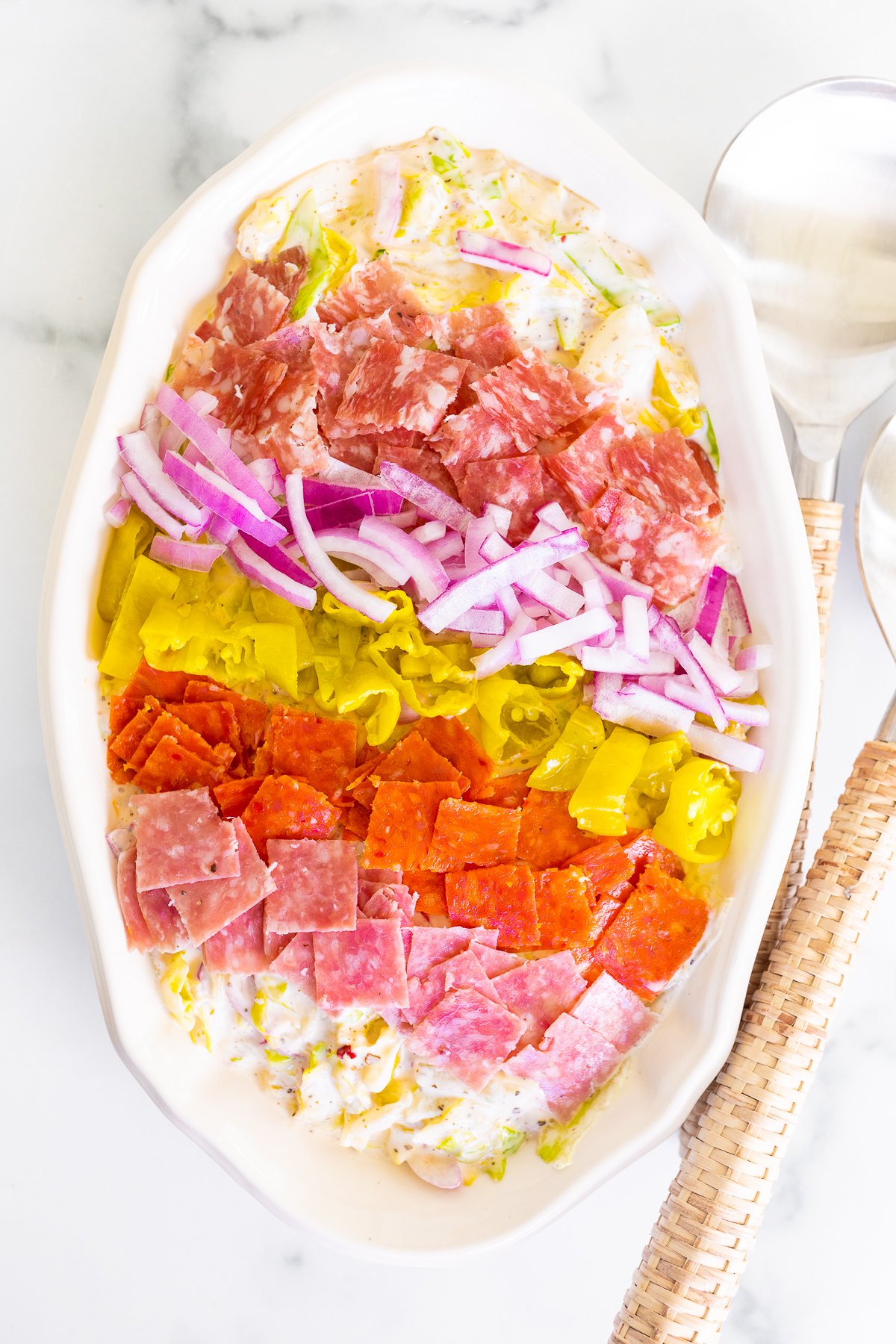 A grinder salad recipe in a white oval bowl, with rattan serving utensils to the side.