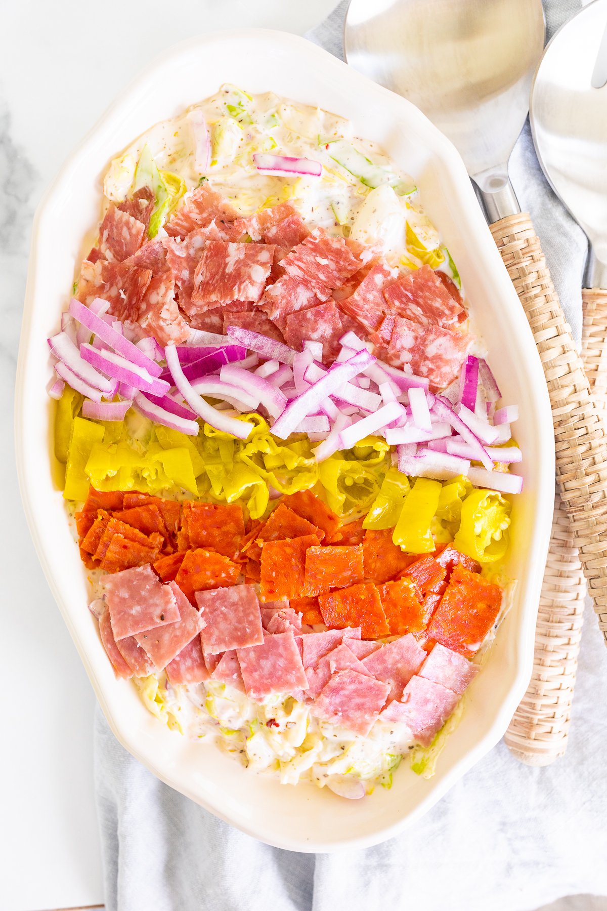 A grinder salad recipe in a white oval bowl, with rattan serving utensils to the side.