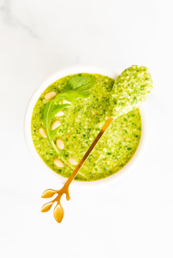 A white bowl full of arugula pesto on a white countertop, gold spoon across the top of the bowl.