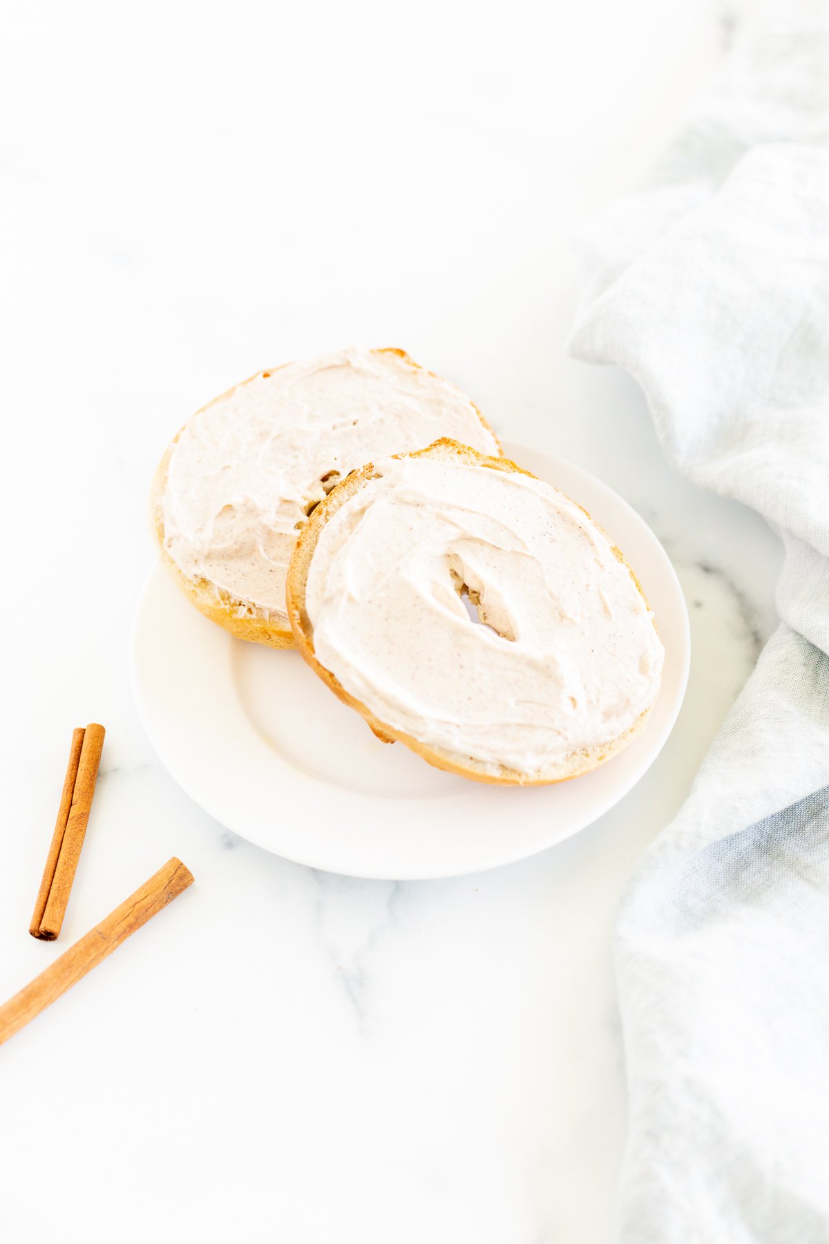 A bagel with cinnamon cream cheese and cinnamon sticks on a white plate.
