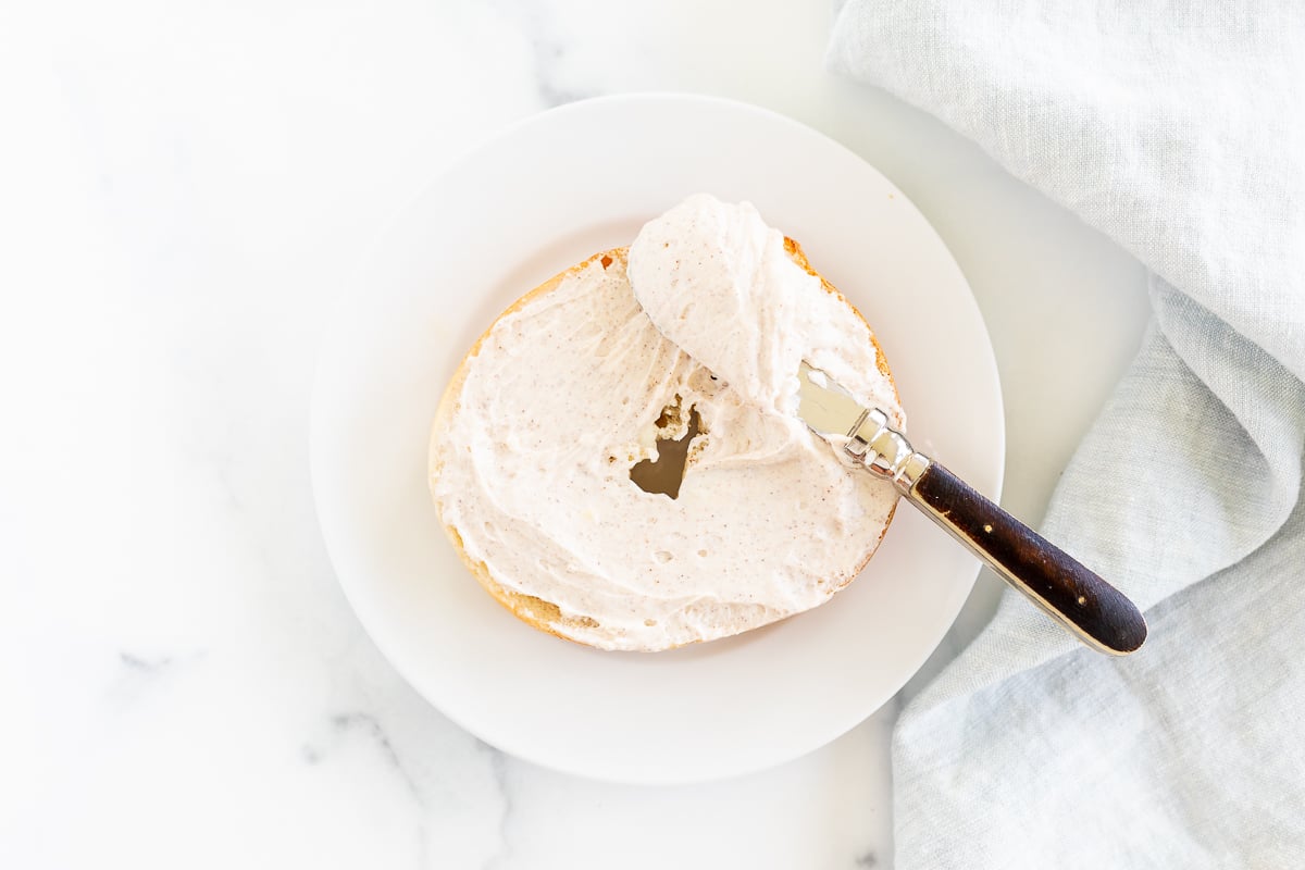 A bagel with cinnamon cream cheese and cinnamon sticks on a white plate.