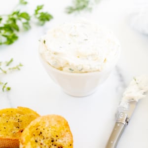 A white bowl of garlic and herb cream cheese on a marble countertop.