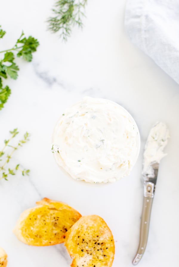 A white bowl of garlic and herb cream cheese on a marble countertop. Fresh herbs nearby.