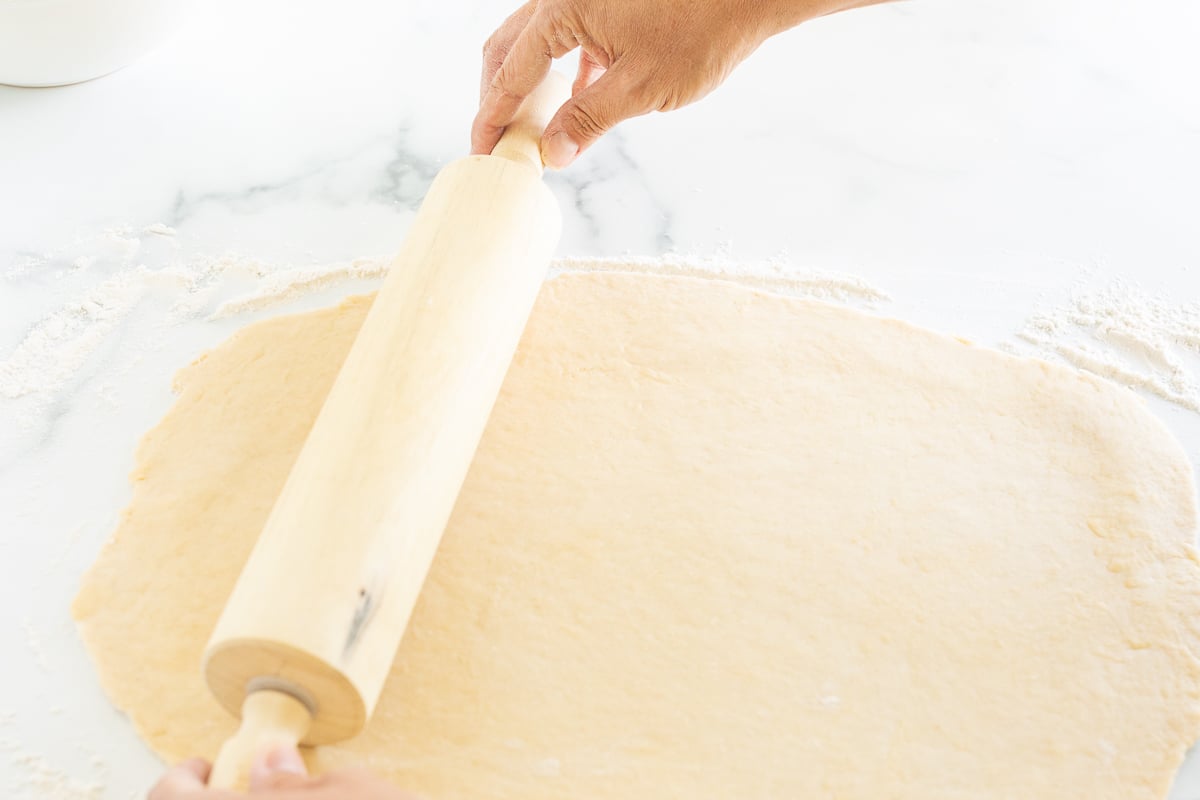 A person preparing apple cinnamon rolls, rolling out dough on a marble countertop.