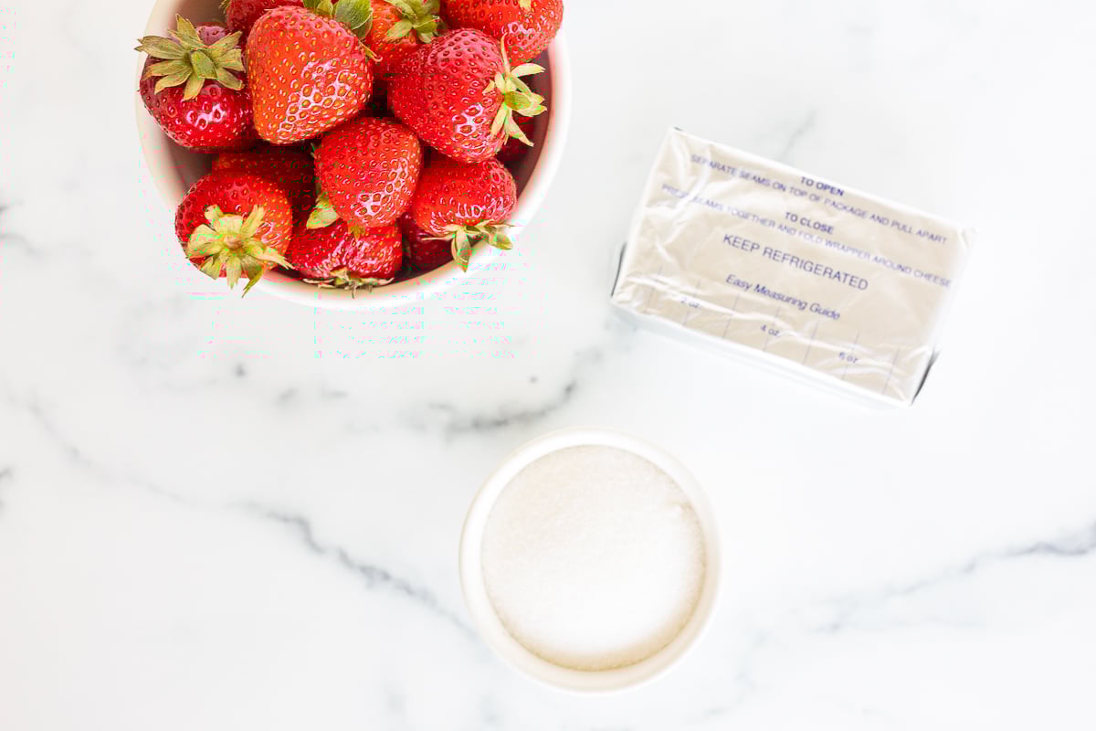 A bowl of strawberries next to a bowl of sugar and block of cream cheese to make strawberry cream cheese.