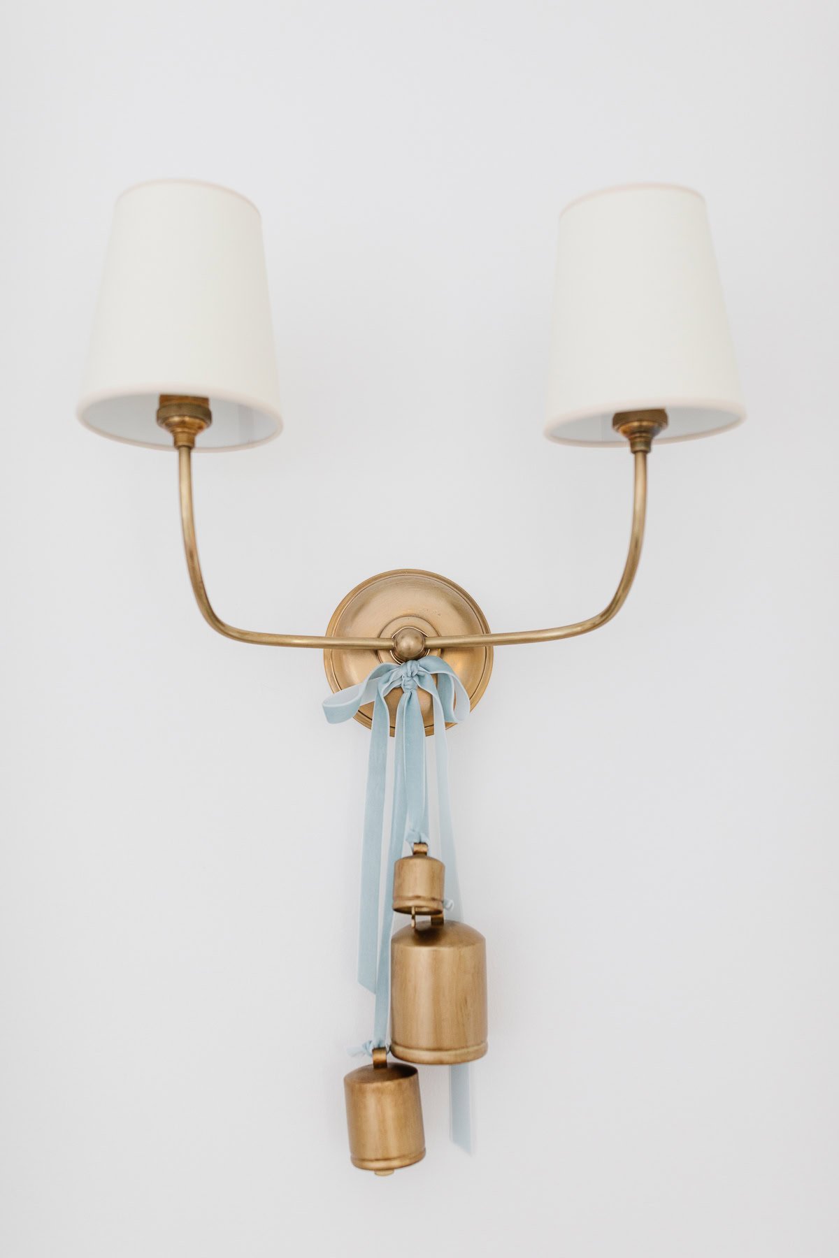 A brass wall sconce with two white lampshades, decorated with blue ribbon and finished with three hanging brass bells for a charming touch.