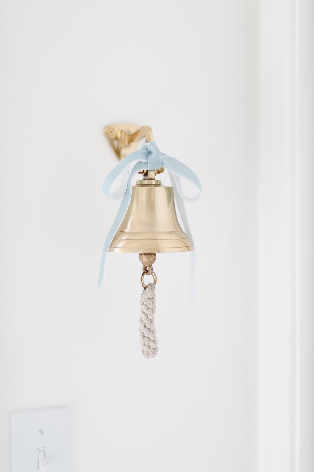 A pair of brass bells with a blue ribbon and braided pull cord is mounted on a white wall above a light switch.