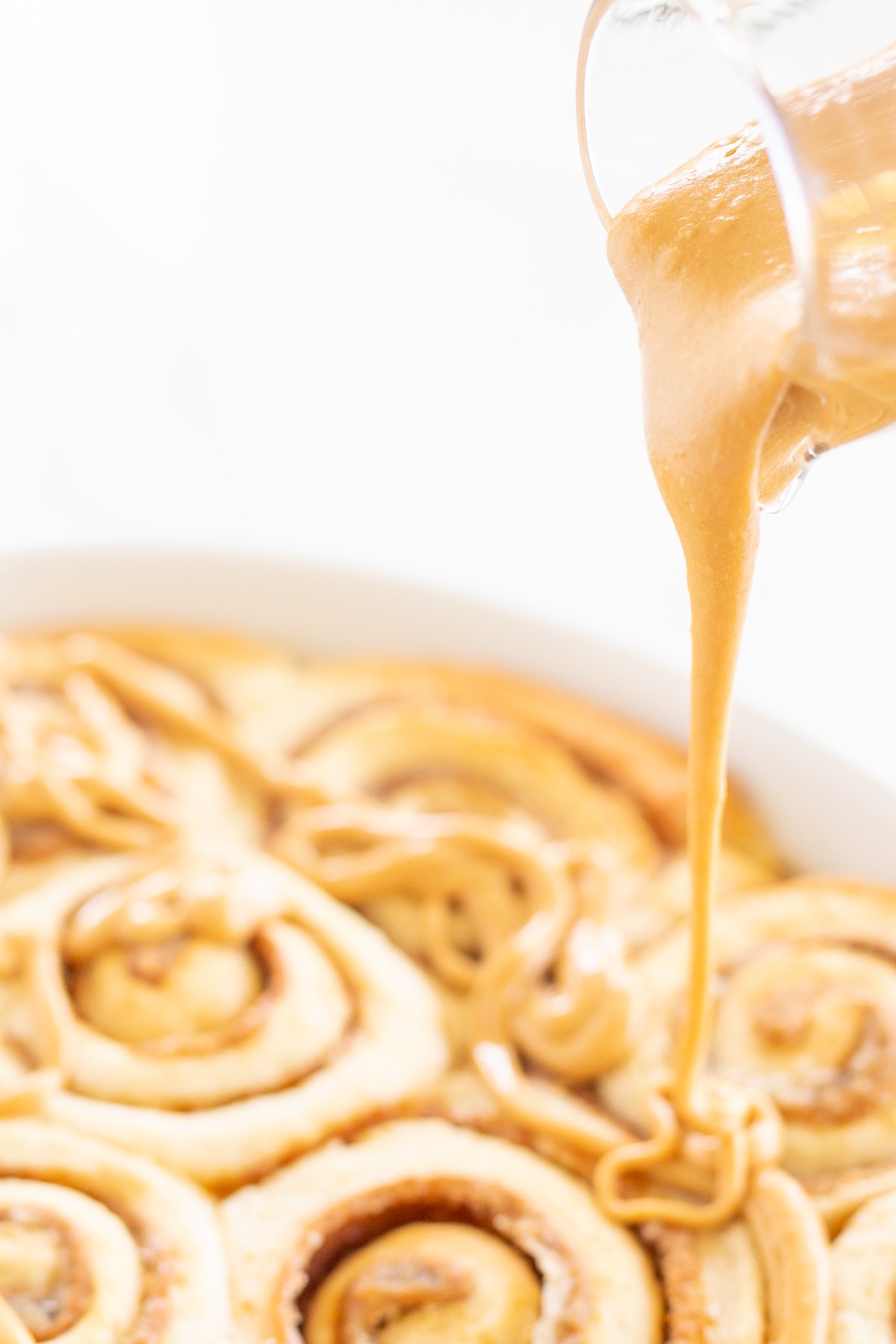 A clear glass pitcher pouring cookie butter frosting over a pan of cinnamon rolls.