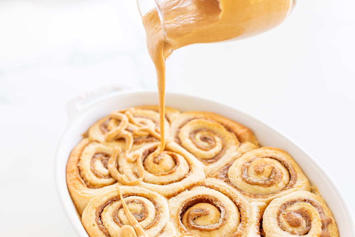 A clear glass pitcher pouring cookie butter frosting over a pan of cinnamon rolls.