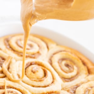 A clear glass pitcher pouring cookie butter frosting over a pan of cinnamon rolls.