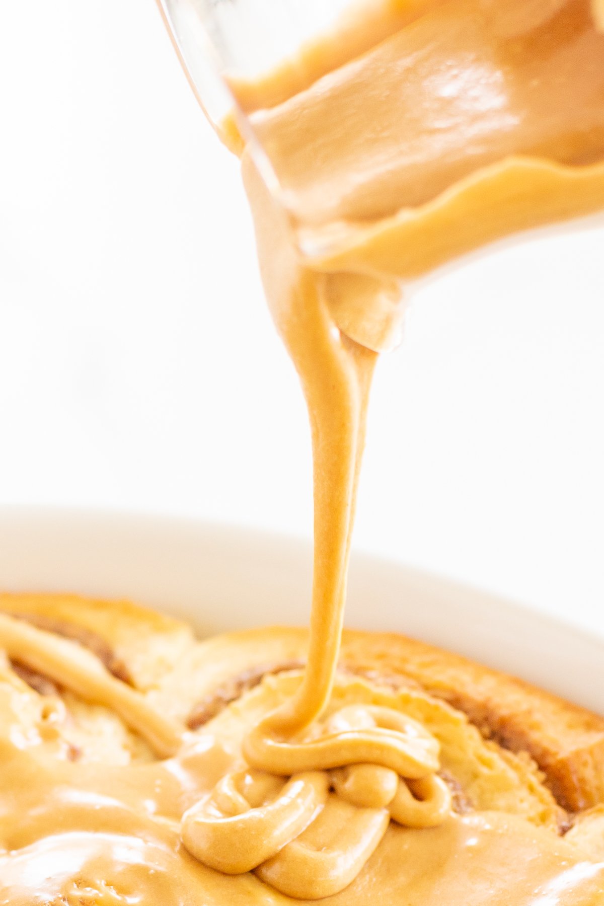 A clear glass pitcher pouring cookie butter frosting over a pan of cinnamon rolls.